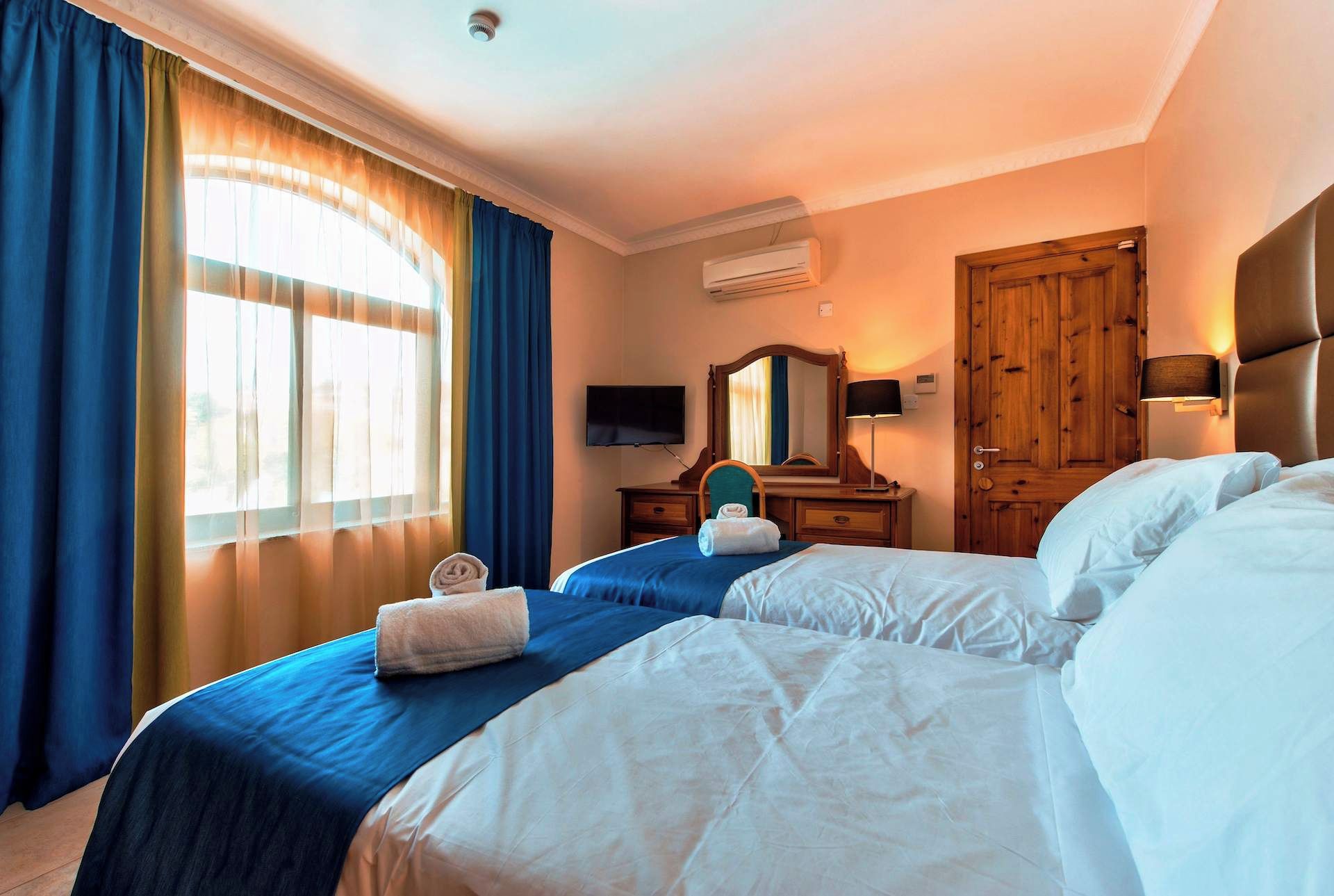 A hotel room with two beds, a desk with a chair, a mirror, an air conditioning unit, and a wooden door.
