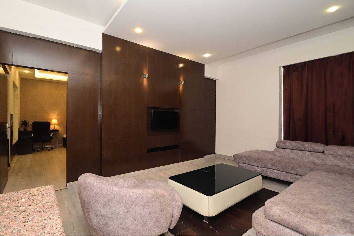Un salon moderne avec un canapé d’angle, une table basse, un téléviseur à écran plat fixé à un mur foncé et un éclairage encastré au plafond.