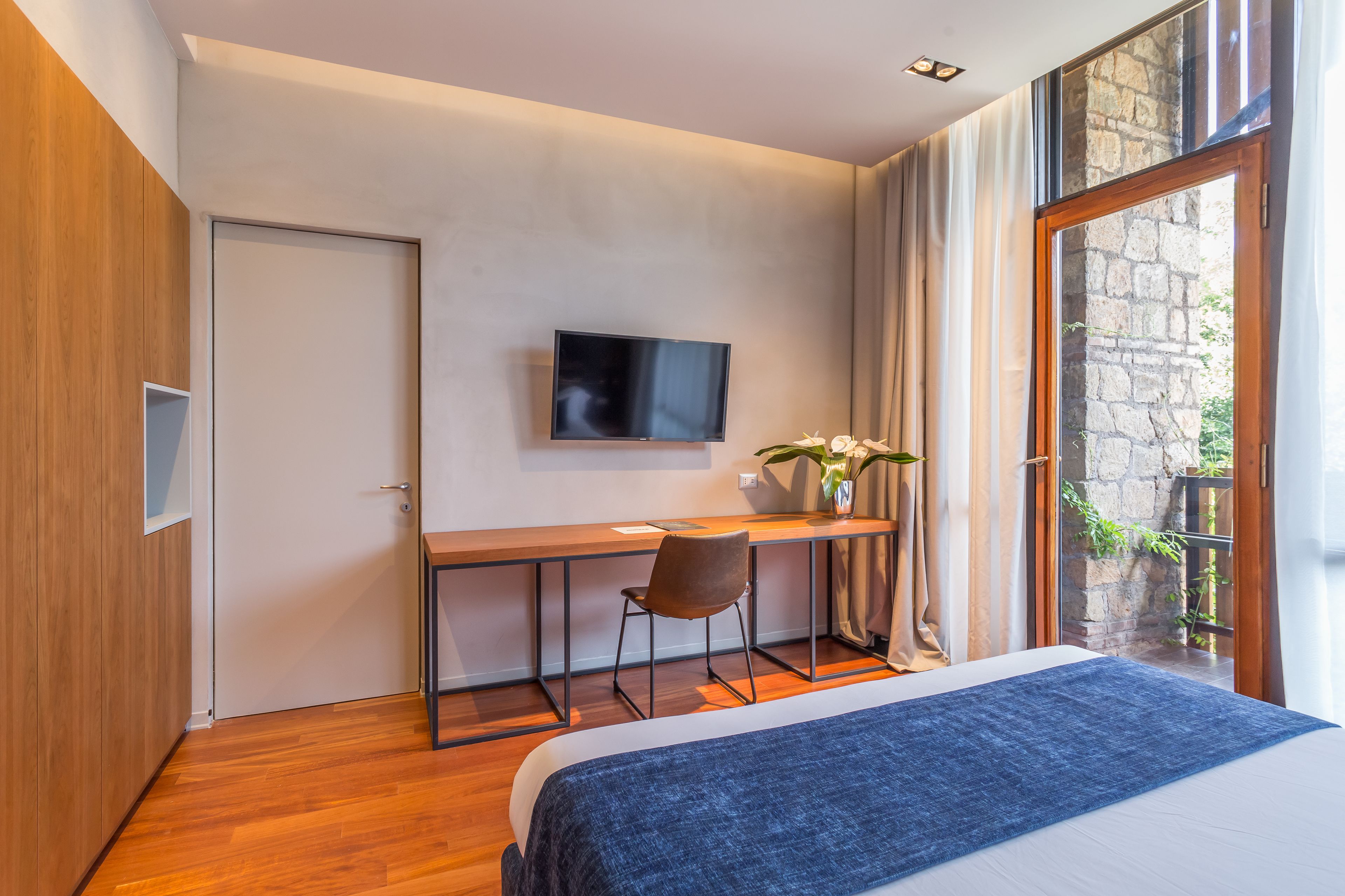 A modern hotel room with a wooden floor, a bed with a blue blanket, a desk with a chair, a flat-screen TV, and a large window with a view of greenery.