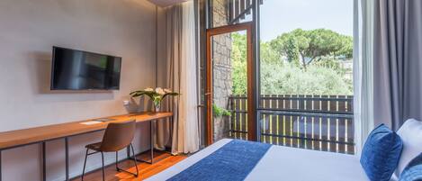 A modern hotel room with a large bed, a desk with a chair, a flat-screen TV, and a balcony with a view of greenery.