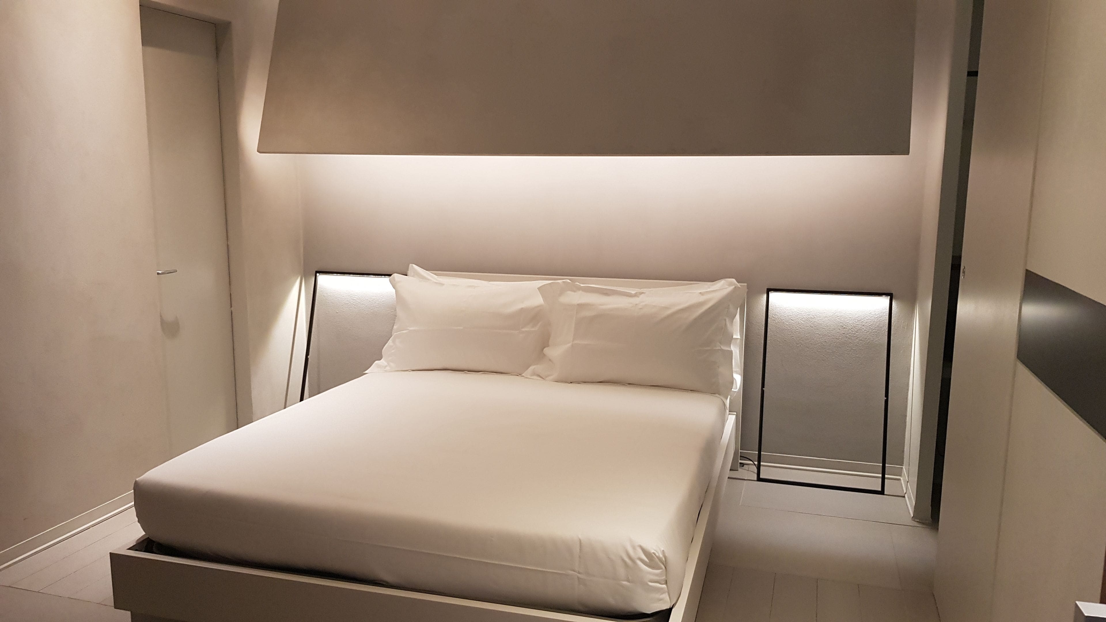 A modern hotel room with a bed, a large window, and a wall-mounted light fixture.