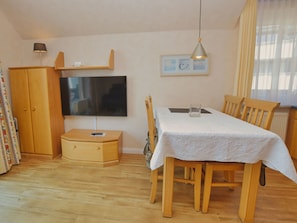 A dining area with a table set for two, a television mounted on the wall, and a cabinet with a remote control on top.