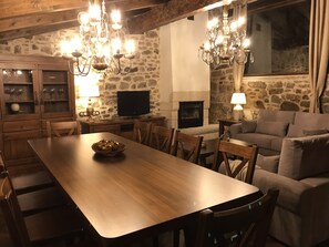 A cozy dining area with a wooden table, chairs, and a chandelier. There is a stone wall, a fireplace, and a television.