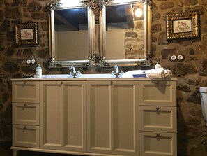 A bathroom with a double sink, two mirrors, and a stone wall. There are two framed pictures on the wall and a towel on the sink.