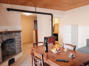 Un comedor con chimenea de piedra, mesa de madera, sillas, una botella de vino, una copa y un plato con fruta.