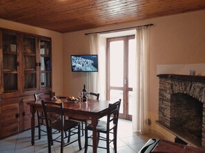 Zona pranzo con tavolo e sedie in legno, televisore montato a parete e camino in pietra.