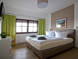 Ein Schlafzimmer mit Bett, Schreibtisch, Stuhl, Heizkörper, einem Fenster mit Vorhängen, einem Bild an der Wand und einer Lampe.
