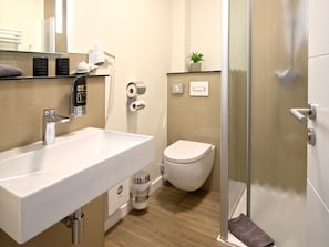 A modern bathroom with a white sink, toilet, and shower. There is a towel rack, a plant, and a hand dryer.