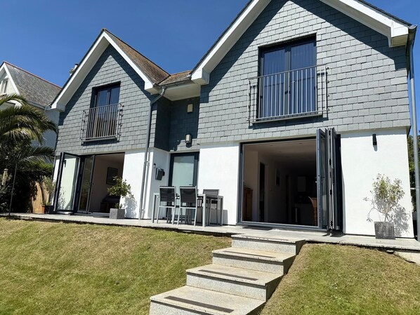 A two-story house with a grey and white exterior, a balcony on the upper floor, and a front door leading to a covered patio area.