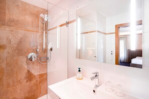 Un baño moderno con cabina de ducha de vidrio, un lavamanos blanco con dispensador de jabón y un espejo grande que refleja un dormitorio.