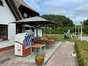 An outdoor seating area with a thatched-roof building, wooden furniture, and a large umbrella providing shade.