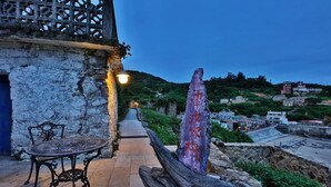 Un bâtiment en pierre avec une table et un banc en métal, une sculpture en bois et un sentier menant à une colline où se trouvent d’autres bâtiments.