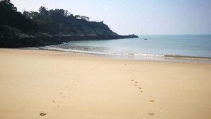 Une plage de sable où l’on peut voir des empreintes de pas menant vers l’eau, une falaise rocheuse et, au loin, un petit bateau.