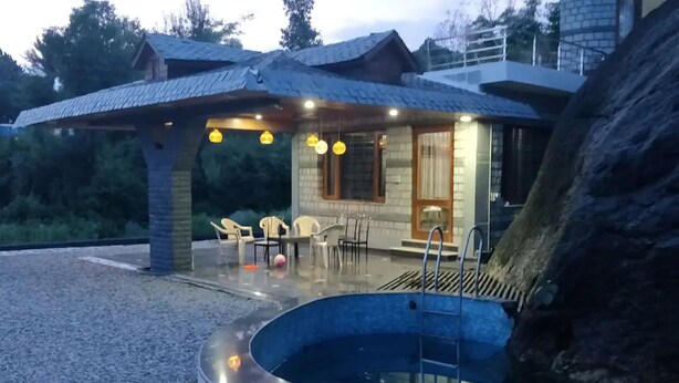 Un área de piscina con una estructura de piedra, asientos y un edificio de madera al fondo.