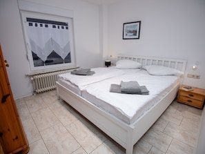 A bedroom with a white bed, a radiator, a window with a view of mountains, a bedside table with a lamp, and a framed picture on the wall.