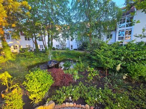 Ein Garten mit Teich, verschiedenen Pflanzen und einem Gebäude im Hintergrund.