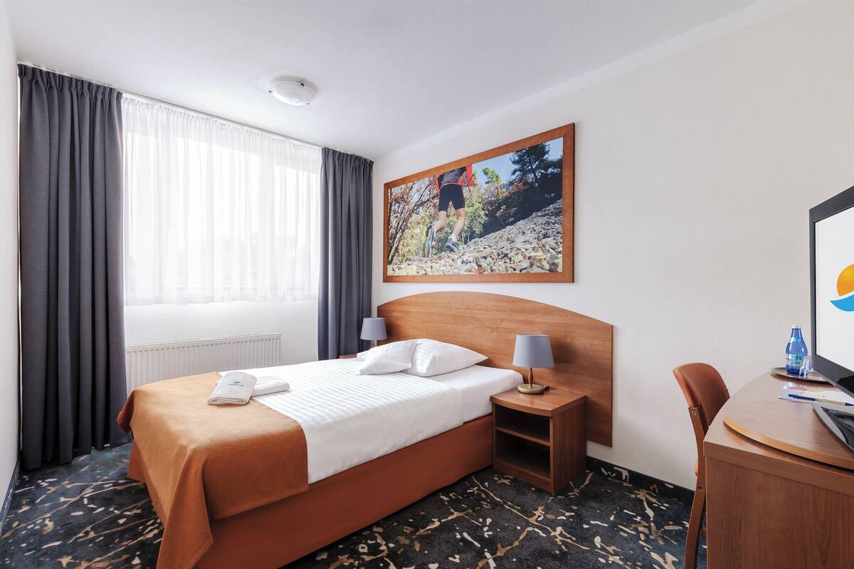 Ein Hotelzimmer mit einem Bett, einem Schreibtisch, einem Stuhl, einem Gemälde und einem Fenster mit Vorhängen.