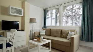 Un salon avec un canapé beige, une table basse blanche, un téléviseur posé sur un meuble blanc et une fenêtre avec des rideaux.