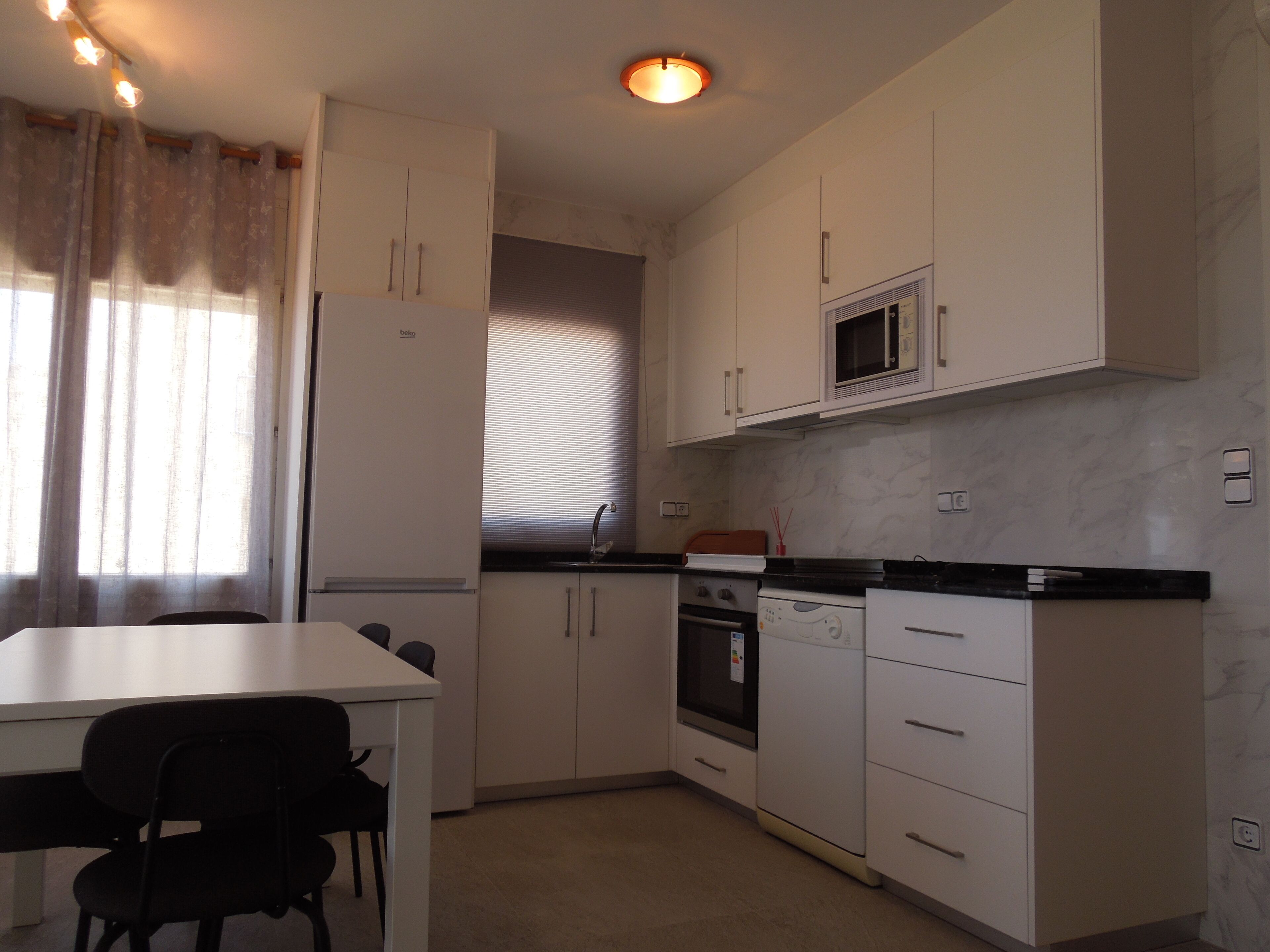 A compact kitchen with white cabinets, a microwave, a stove, and a sink. There is a dining table with chairs and a window with curtains.