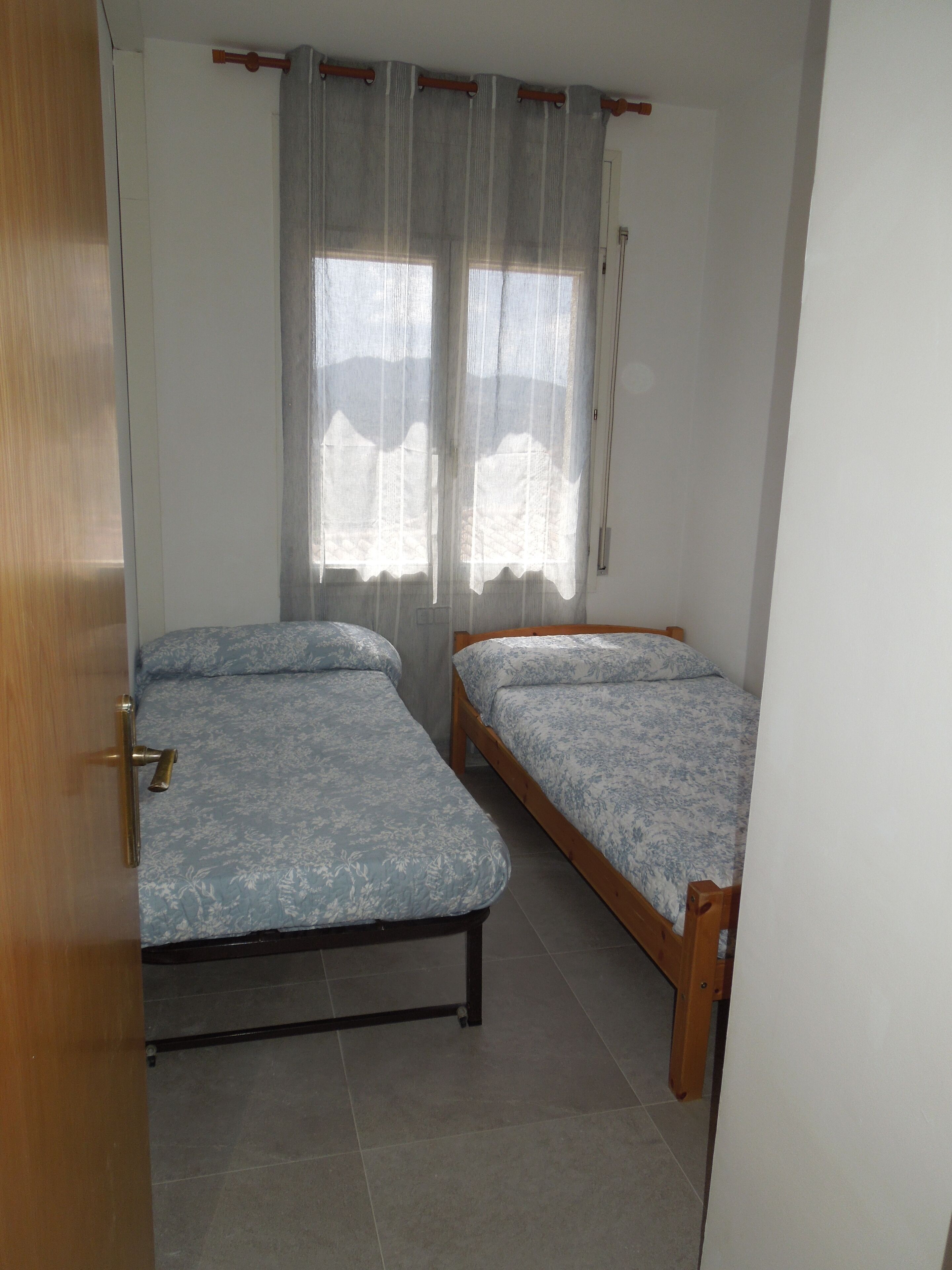 Quarto com duas camas de solteiro, porta de madeira e janela com cortinas leves.
