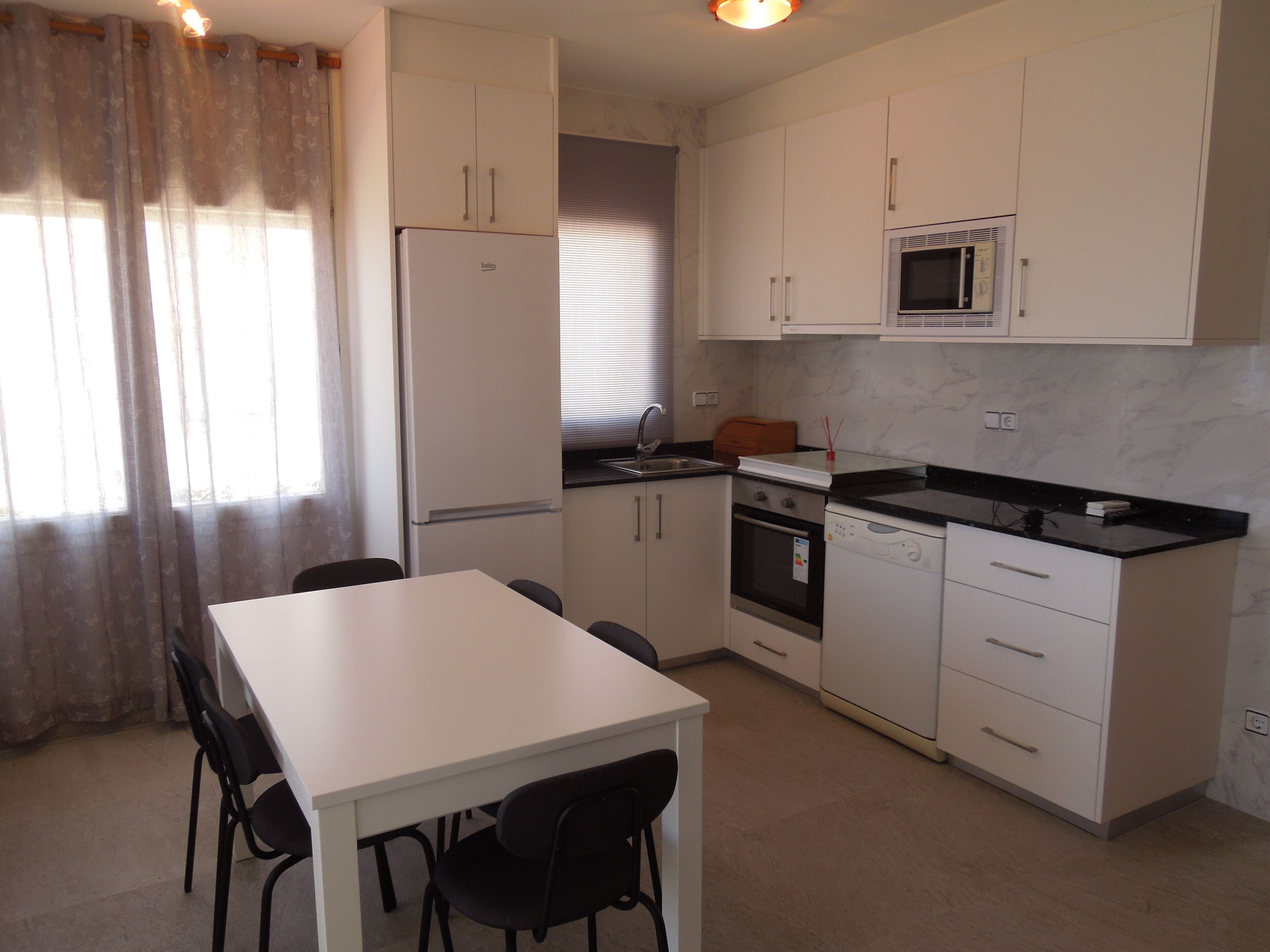 Cocina compacta equipada con refrigerador, microondas, horno y lavavajillas. Incluye una mesa de comedor con sillas y una ventana con cortinas.