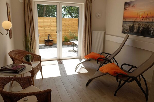 A room with a sliding glass door leading to an outdoor area, wicker furniture, a table with magazines, and a painting of a sunset. - Apartment courtyard - Ferienhof Börsby - Apartment courtyard (Pommerby)