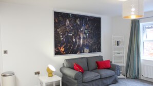 A modern living room with a grey sofa, red cushions, a white side table with a lamp, a white bookshelf, and a large cityscape painting on the wall. - Fabulous Apartment in the City of Ripon, close to Cathedral with private parking (Ripon)