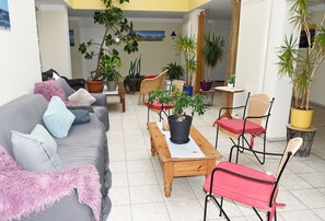 Un salon avec un canapé gris, une table basse en bois et plusieurs fauteuils. On y trouve de nombreuses plantes en pot et une vue sur l’extérieur.