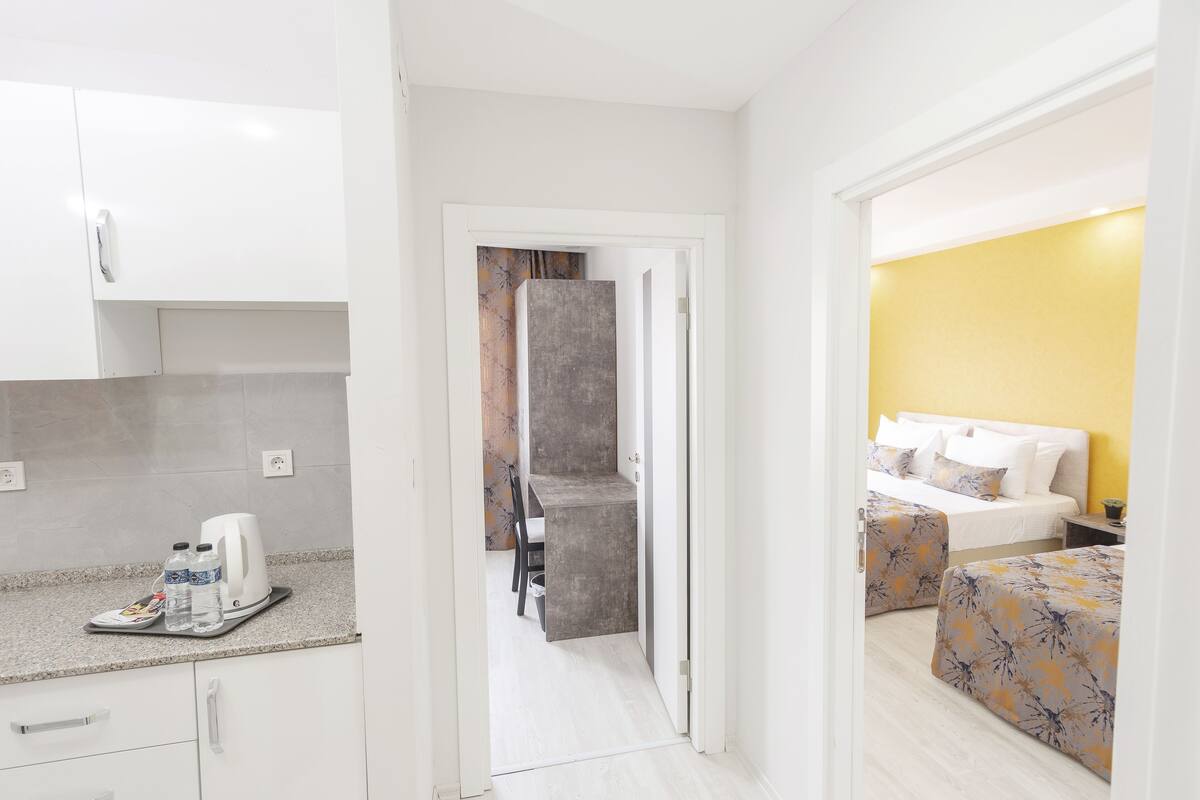 Une chambre d’hôtel avec une kitchenette, un lit recouvert d’une literie à motifs floraux et donnant sur le couloir.