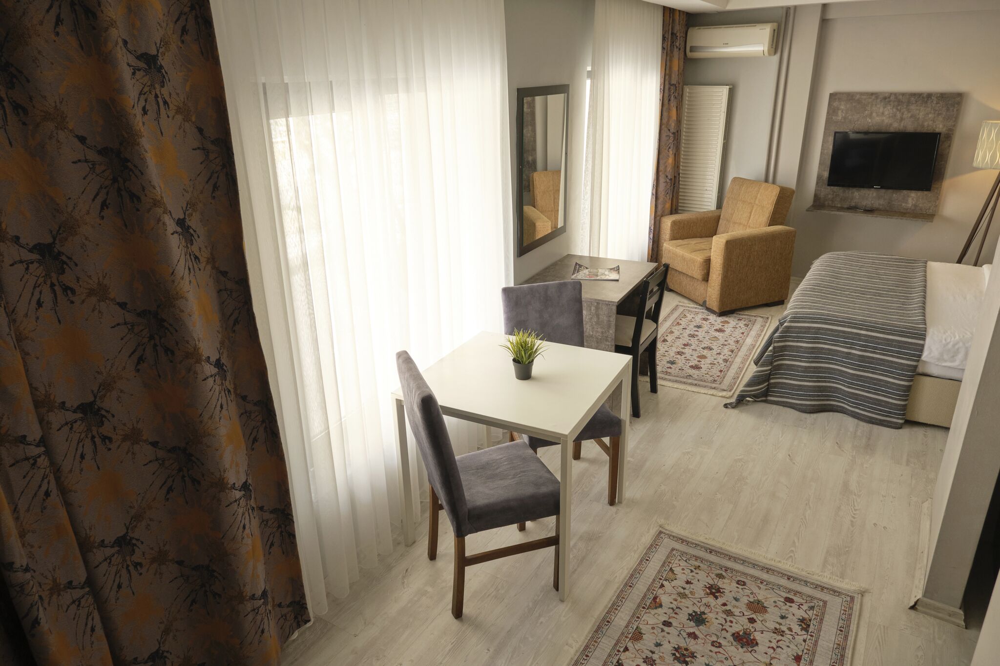 A hotel room with a bed, a sofa, a dining table with two chairs, a small table with a plant, a mirror, a TV mounted on the wall, and a window with curtains.