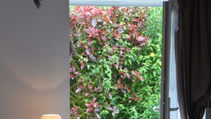 Een slaapkamer met een bed, een nachtkastje met een lamp en uitzicht op een tuin door een open deur.