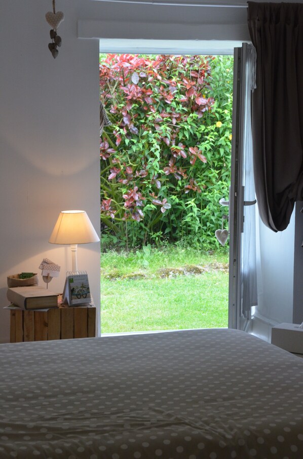 A bedroom with a bed, a bedside table with a lamp, and a view of a garden through an open door. - Le Petit Monty (Saint-Malô-du-Bois)