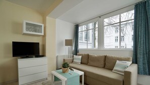 Een woonkamer met een televisie op een witte dressoir, een beige bank met gestreepte kussens, een tafeltje met een plant en een raam met gordijnen.
