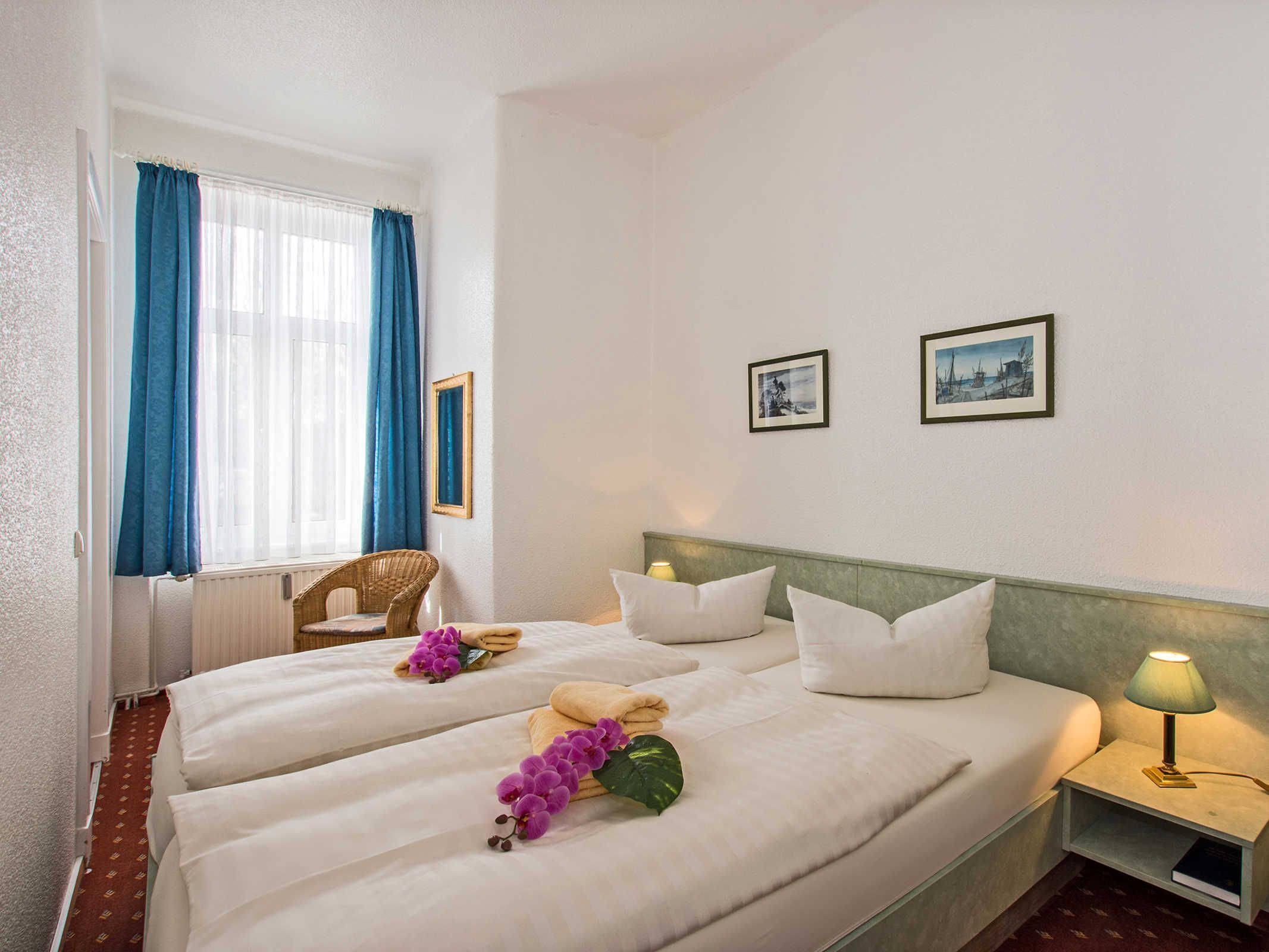 A hotel room with two beds, a chair, a nightstand with a lamp, and two framed pictures on the wall.