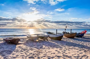 Une plage où plusieurs bateaux sont amarrés sur le sable, un ciel dégagé et le soleil se couche sur l’océan.