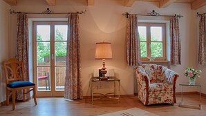 Een kamer met houten balken in het plafond, een bloemen fauteuil, een houten stoel, een tafel met een lamp en een uitzicht door grote ramen.