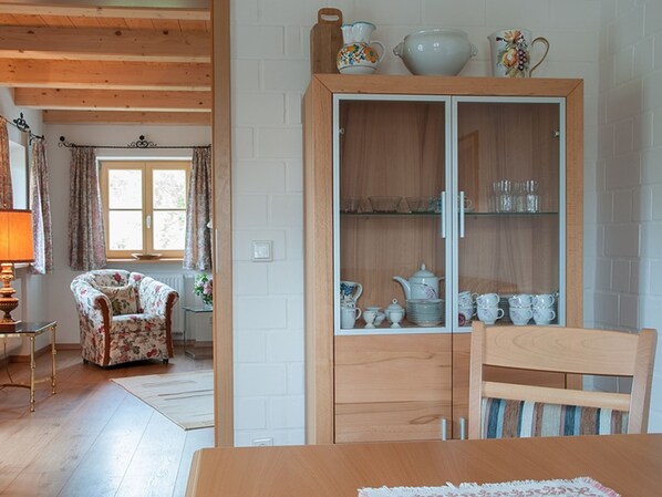 Una mesa de comedor de madera con una silla estampada con motivos florales, un aparador con puertas de cristal que exhibe vajilla y una ventana con cortinas.