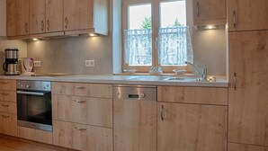 Een keuken met houten kastjes, een ingebouwde oven, een vaatwasser, een raam met gordijnen en een klok aan de muur.