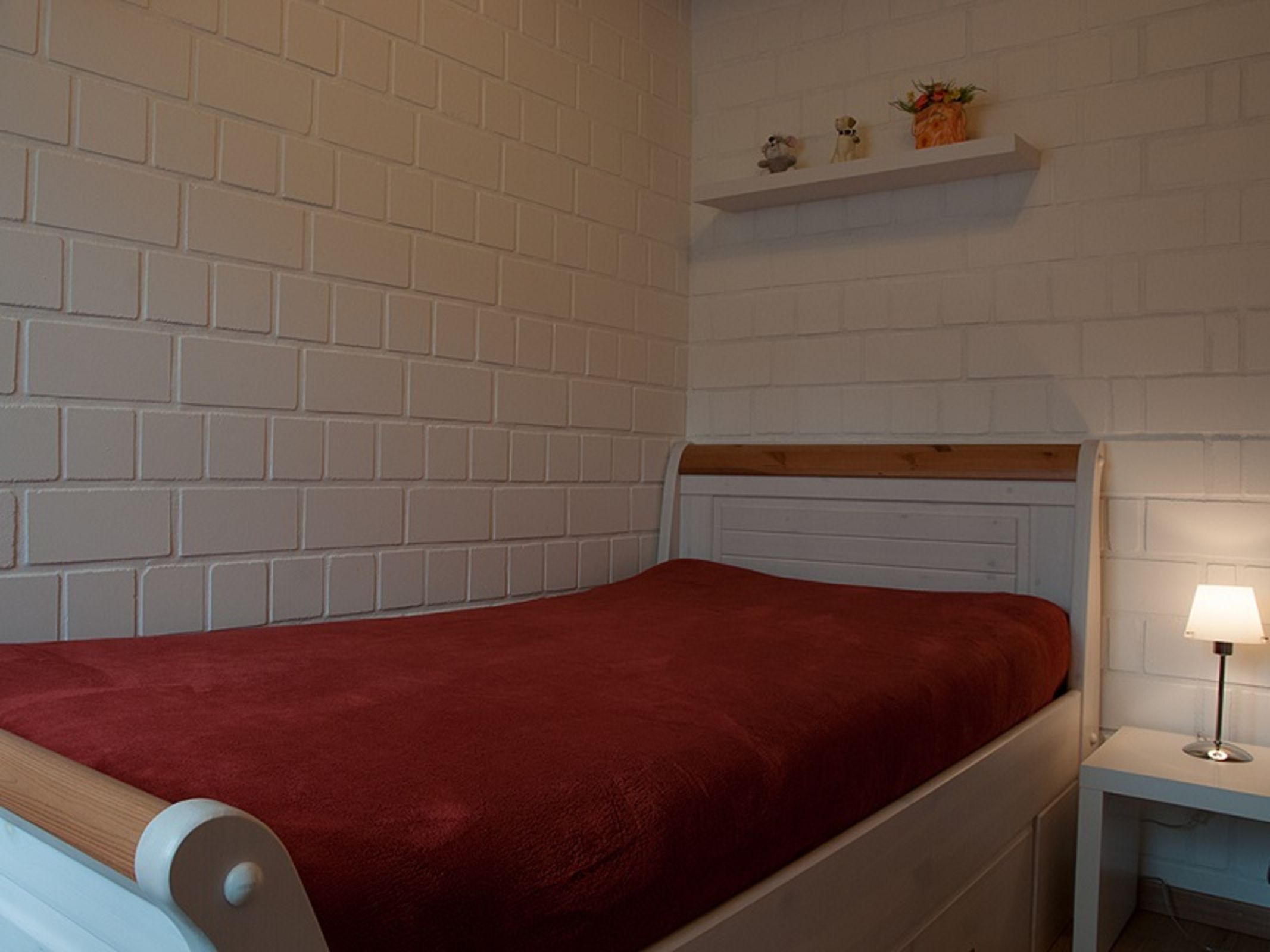 Quarto com parede de tijolos brancos, uma cama de solteiro com cabeceira de madeira, colcha vermelha, mesa de cabeceira com abajur e prateleira com objetos decorativos.