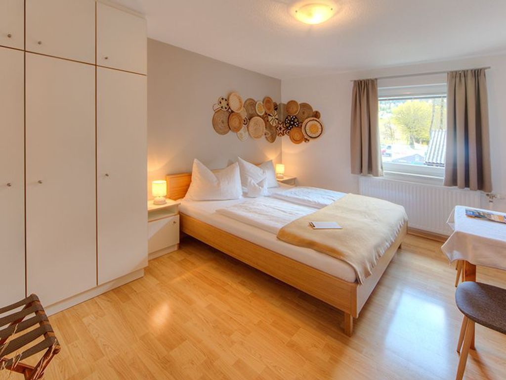 Ein Schlafzimmer mit Bett, Nachttischen, einem Kleiderschrank, einem Wandbild, einem Fenster mit Vorhängen und einem kleinen Tisch mit Stuhl.