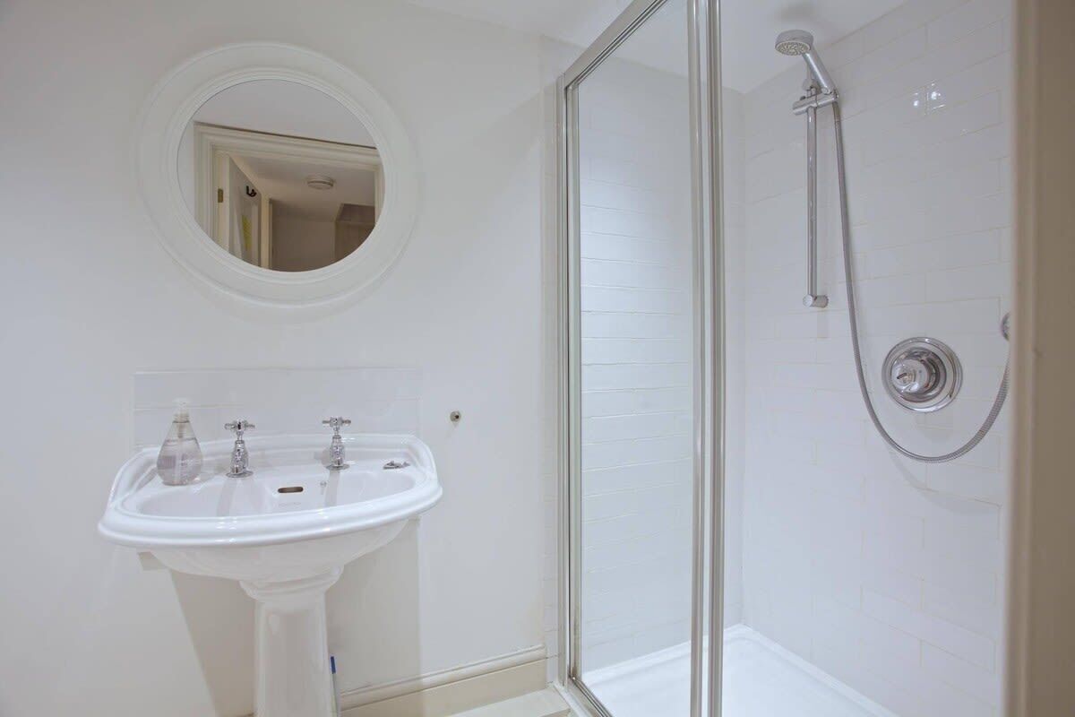 A bathroom with a white pedestal sink, a round mirror, and a glass-enclosed shower.