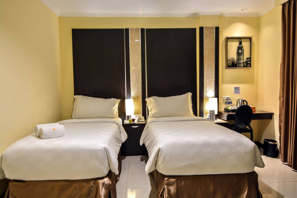 A hotel room with two beds, a desk, a chair, a lamp, and a framed picture on the wall.