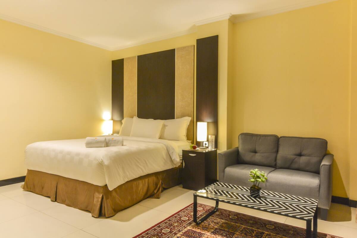 A hotel room with a bed, a gray sofa, a small table with a plant, and a nightstand with a lamp.