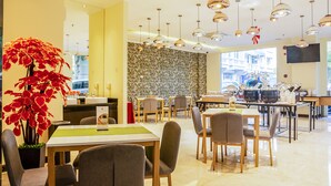A modern dining area with multiple tables and chairs, a large potted plant, and a patterned wall. - Sunbreeze Hotel Senayan (Jakarta)