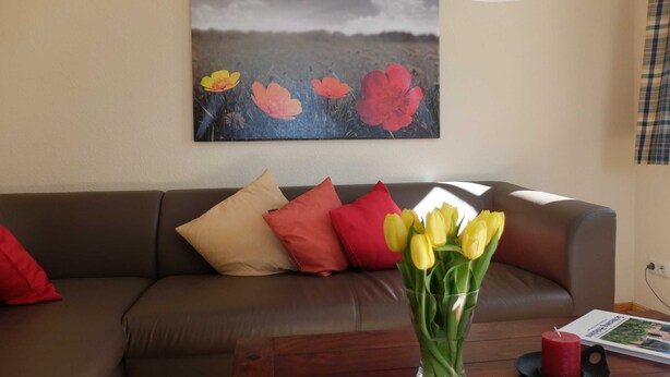 A living room with a brown leather sofa, red and beige cushions, a glass vase with yellow tulips, a red candle, and a magazine on a wooden coffee table. - Apartment Nice view - Ferienhof Börsby - Apartment Schöne Aussicht (Pommerby)