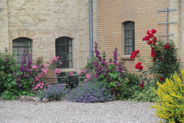 A building with a brick facade, two windows with green frames, and a gravel garden with various flowering plants.