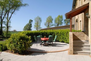Een terras buiten met een tafel en stoelen, omgeven door groen en een bakstenen muur.
