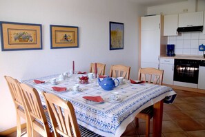A dining area with a table set for four, surrounded by wooden chairs. There are framed pictures on the wall, a kitchen area with white cabinets, and a blue and white patterned tablecloth.