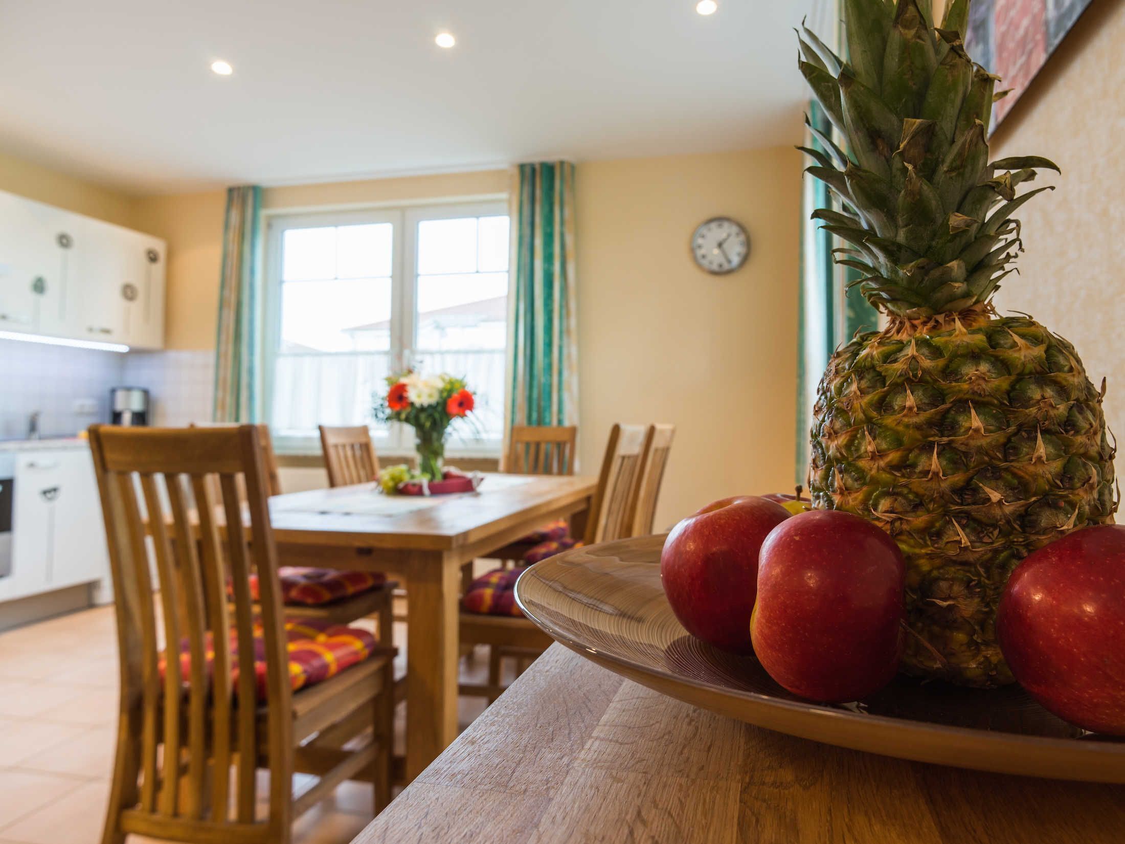 A dining area with a table, chairs, a clock, and a pineapple with apples on a plate.