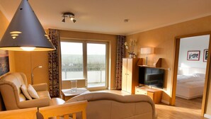 A modern hotel room with a sofa, a dining table, a TV, and a balcony with a view. - N 8K (BC.a5) - Stranddomicil Steiner (Büsum)